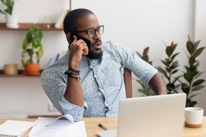 Homme d'affaires afro-américain sérieux parlant au téléphone assis au bureau avec un ordinateur portable, directeur noir concentré faisant un appel d'affaires ayant une conversation mobile sur un téléphone portable contactant le client